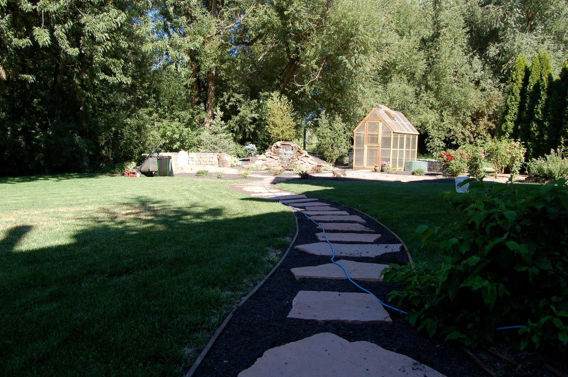 A winding stone pathway through a green lawn, leading toward a stone rock feature and a small wooden shed in a backyard.