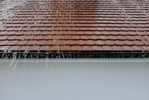 Rainwater flowing from a residential shingle roof during heavy rainfall with visible roof drainage and runoff.