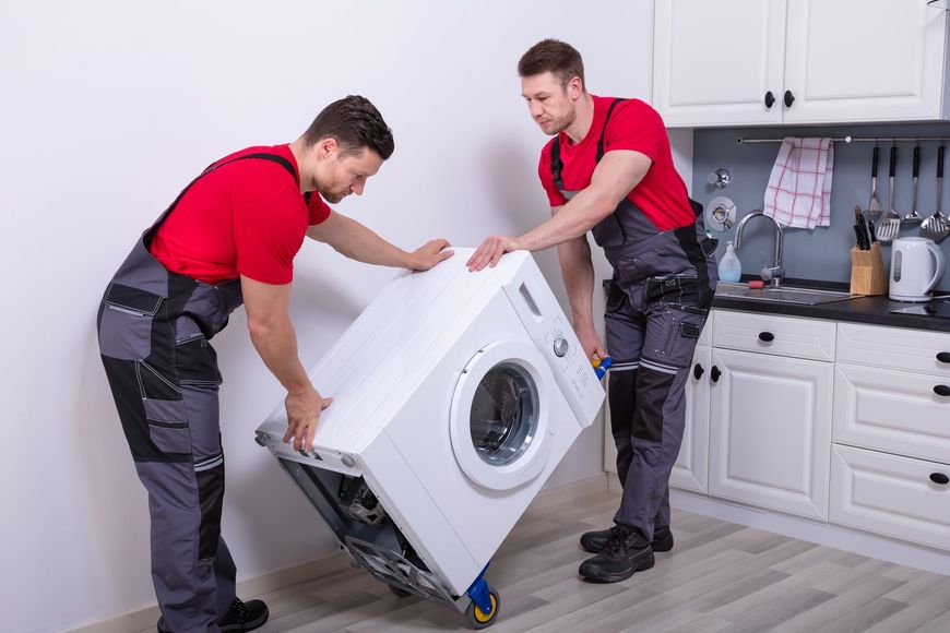 Two people in red shirts and gray overalls moving a washing machine in a kitchen.