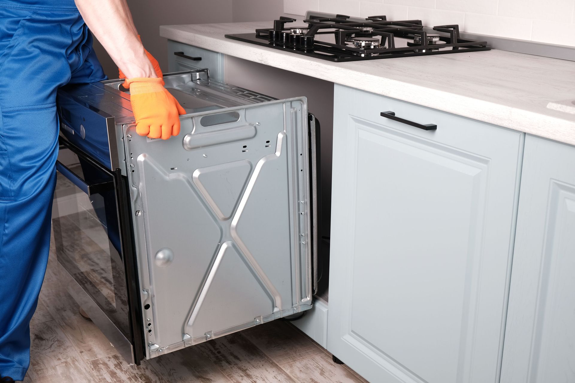 Person installing an oven in a kitchen cabinet, wearing orange gloves and blue overalls.