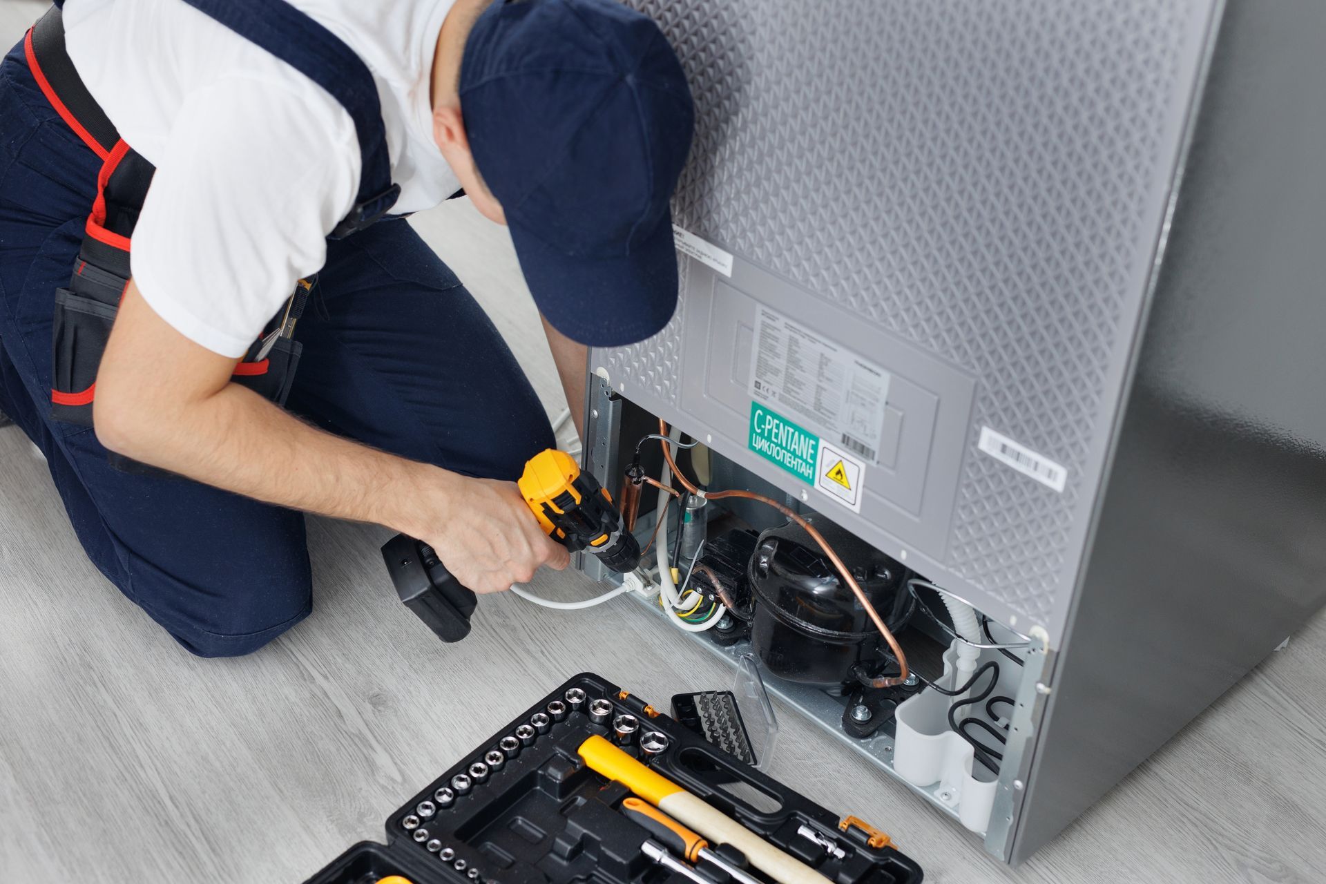 Technician in blue overalls repairs a refrigerator, using a drill. A toolbox sits on the floor.