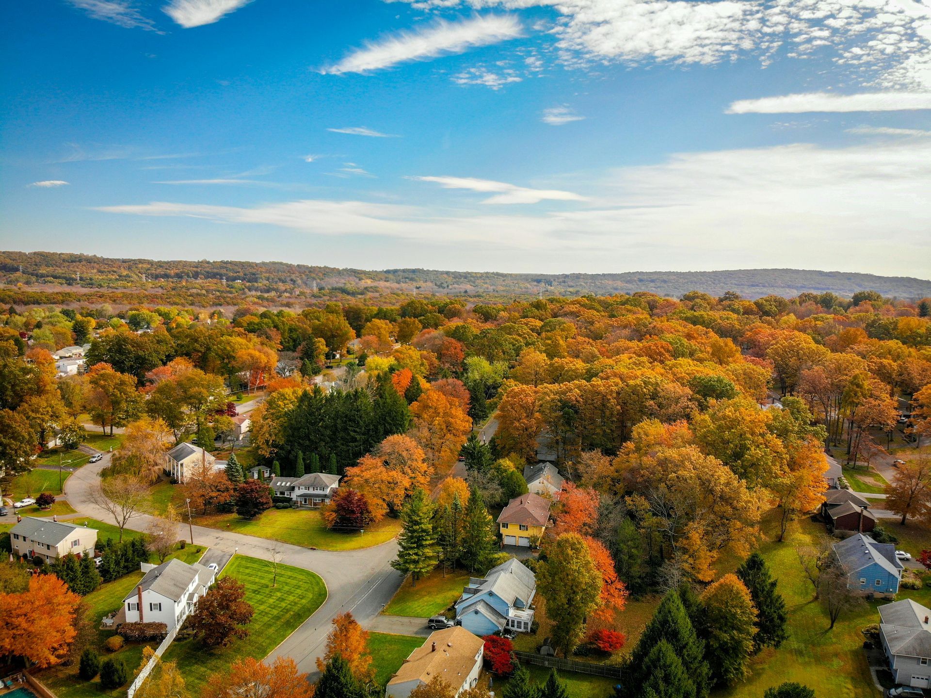 Aerial view of a suburban neighborhood in autumn; trees with yellow, orange, and red leaves, houses with green lawns.