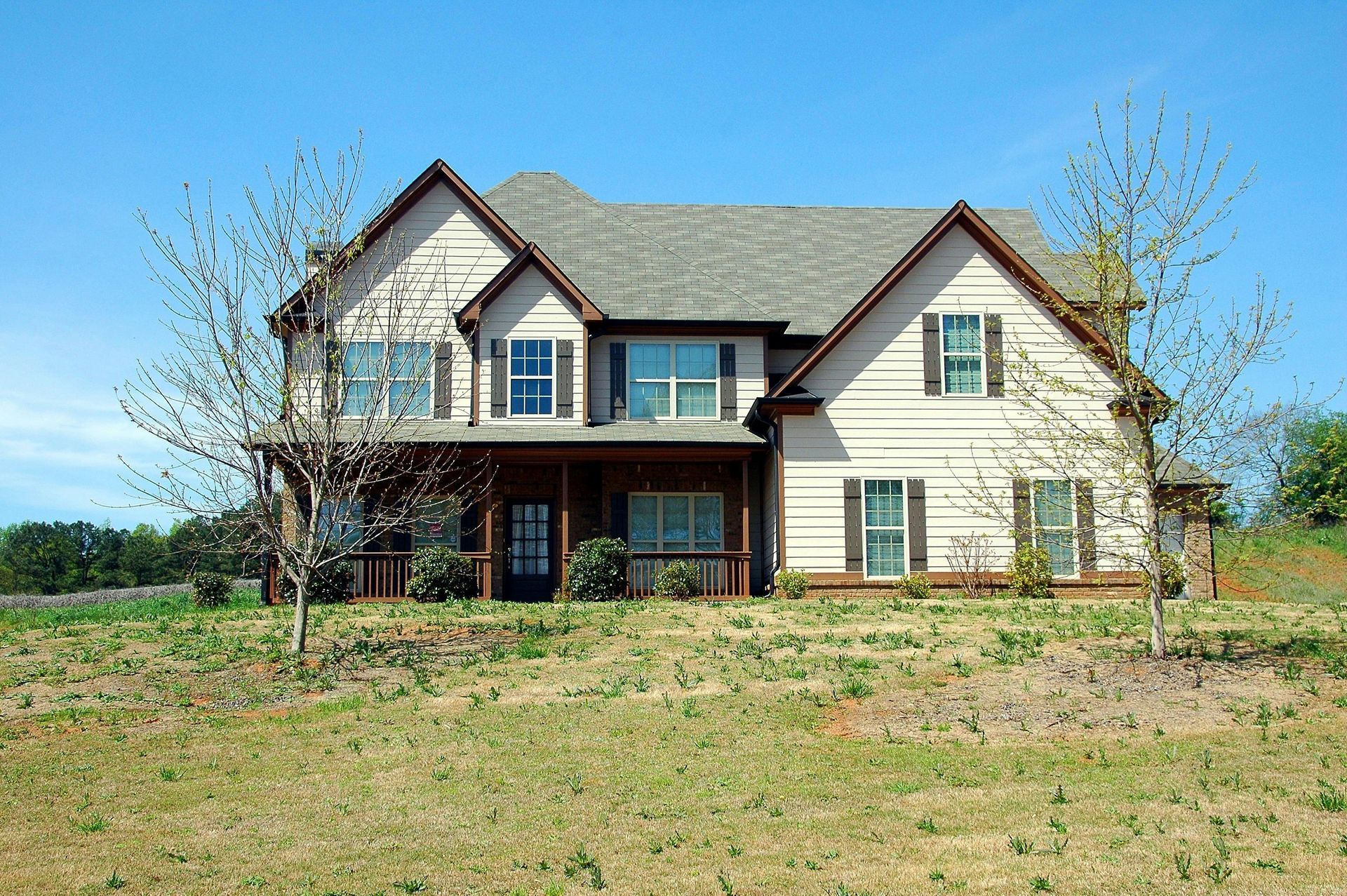 Two-story beige house with brown trim and shutters, sitting on a grassy hill under a blue sky.