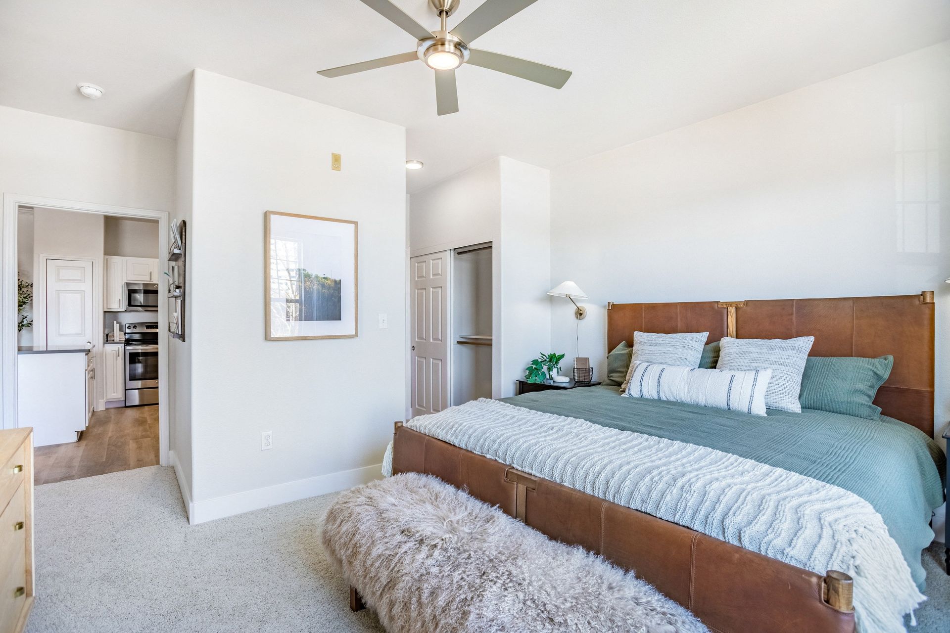 Bedroom with a brown leather bed, green bedding, a furry bench, and a hallway to a kitchen at The Viridian in Greenwood Village, CO.