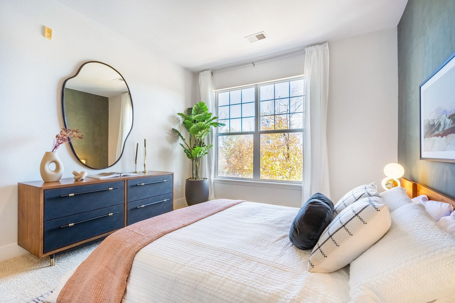 Bedroom with a white bed, blue dresser, large mirror, and a window overlooking trees at The Viridian in Greenwood Village, CO.