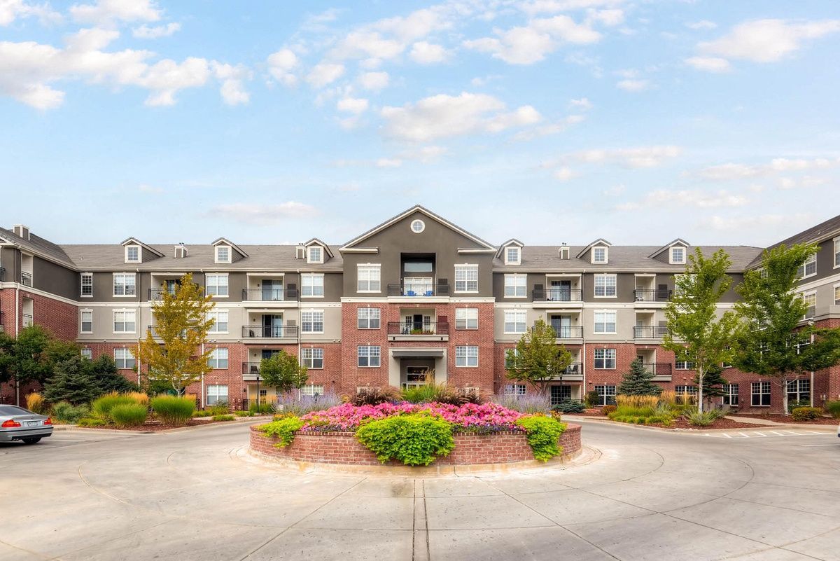 Apartment building with brick and gray siding, flower bed in front, under a blue sky at The Viridian in Greenwood Village, CO.