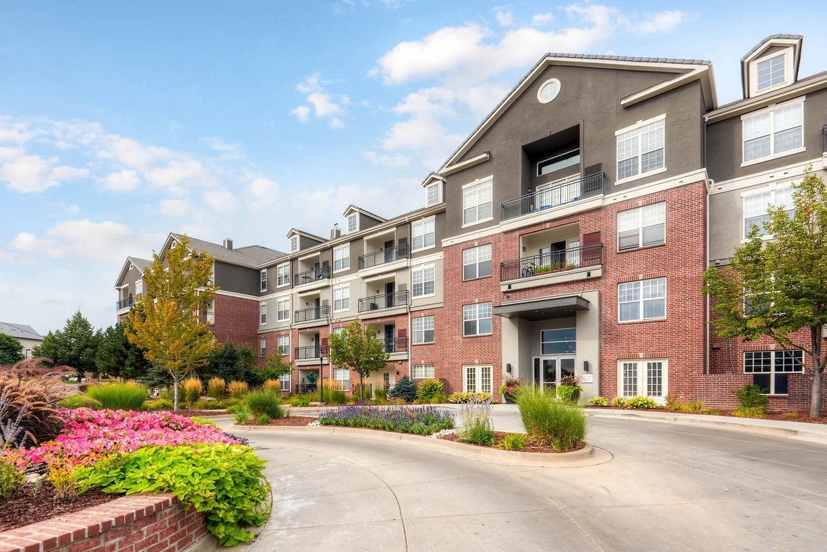 Exterior view of a brick apartment building with a curved driveway, flowers, and a blue sky at The Viridian in Greenwood Village, CO.