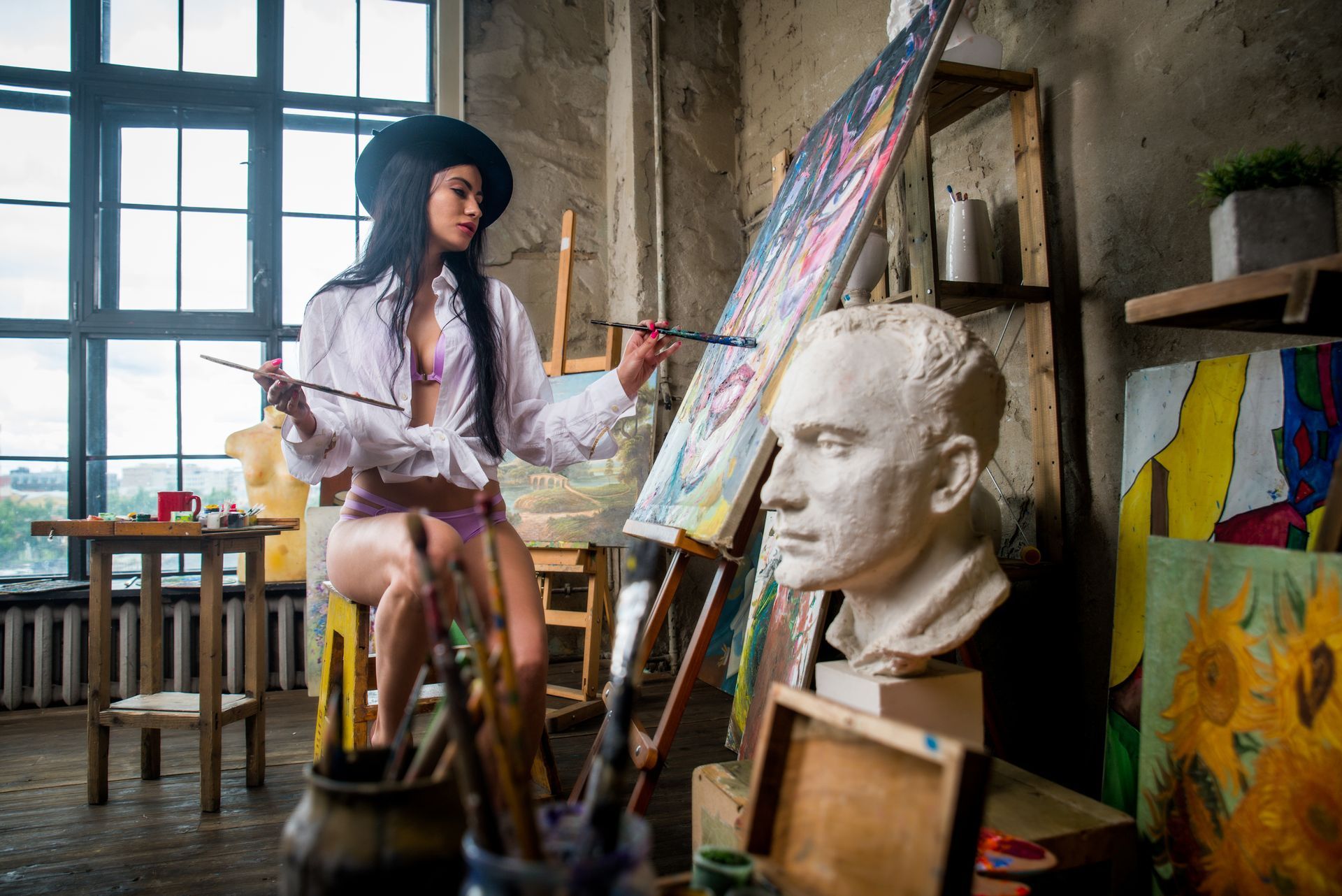 Woman in hat paints at an easel in a studio, with a bust and colorful paintings.
