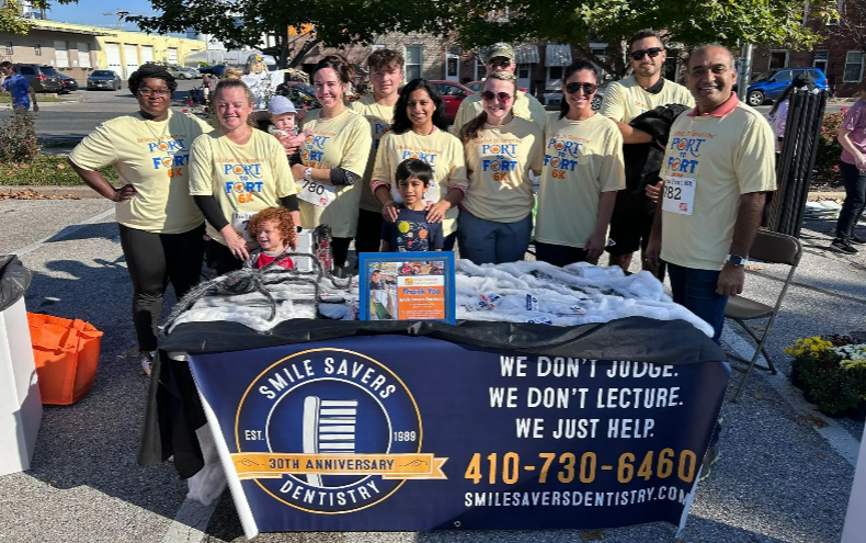 Group of people at a Smile Savers Dentistry event; smiling, outdoors, standing behind a table with supplies.