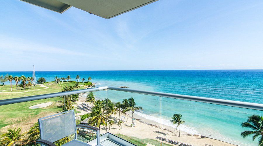 Balcony overlooking beach and turquoise ocean; palm trees and golf course in view.