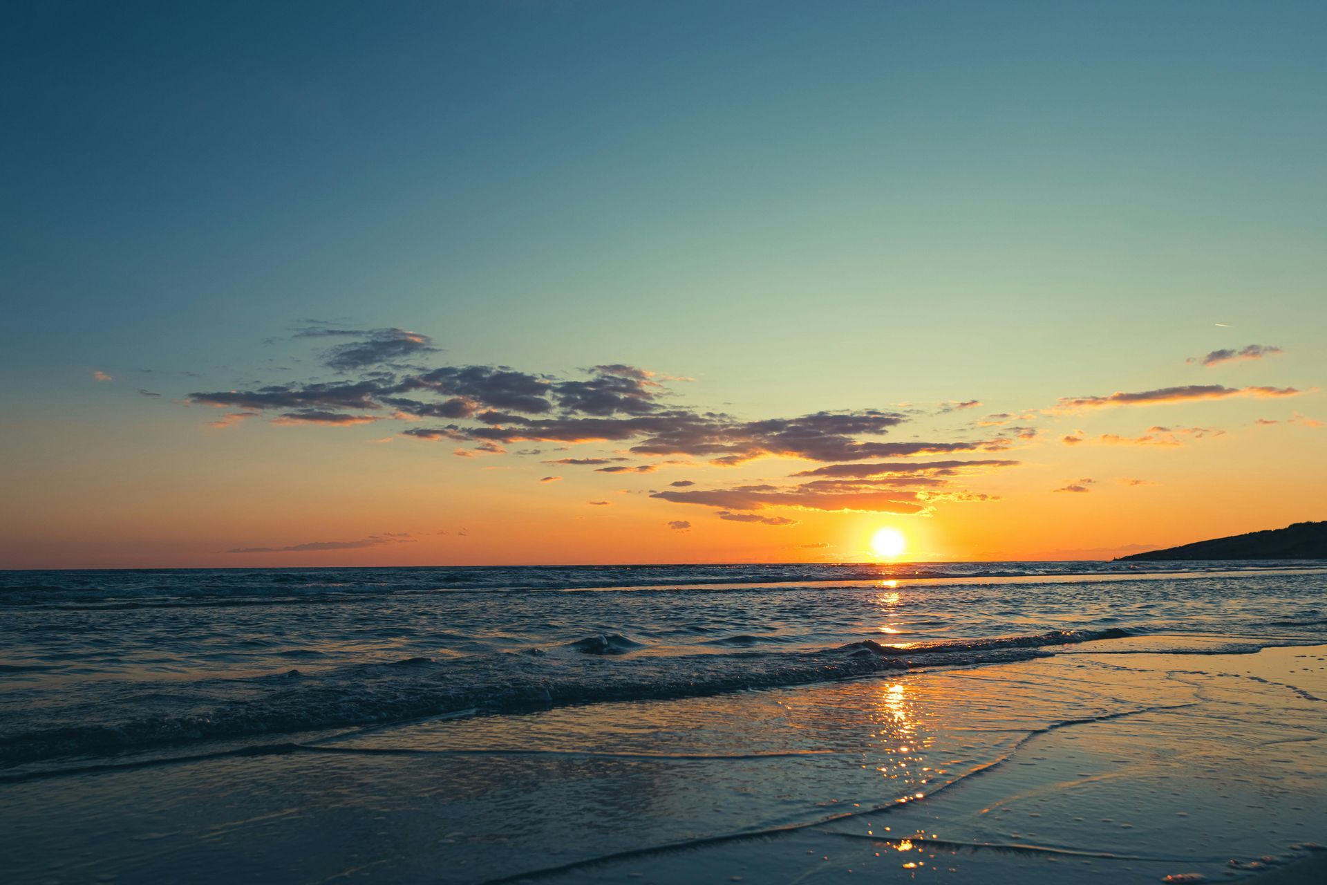 Sunset over the ocean. Orange sun reflecting on wet sand and rippling waves under a blue and orange sky.