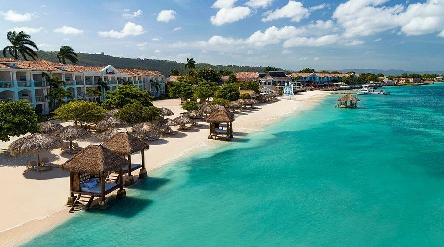 Beach resort with turquoise water, white sand, and thatched-roof cabanas. Buildings and palm trees in the background.