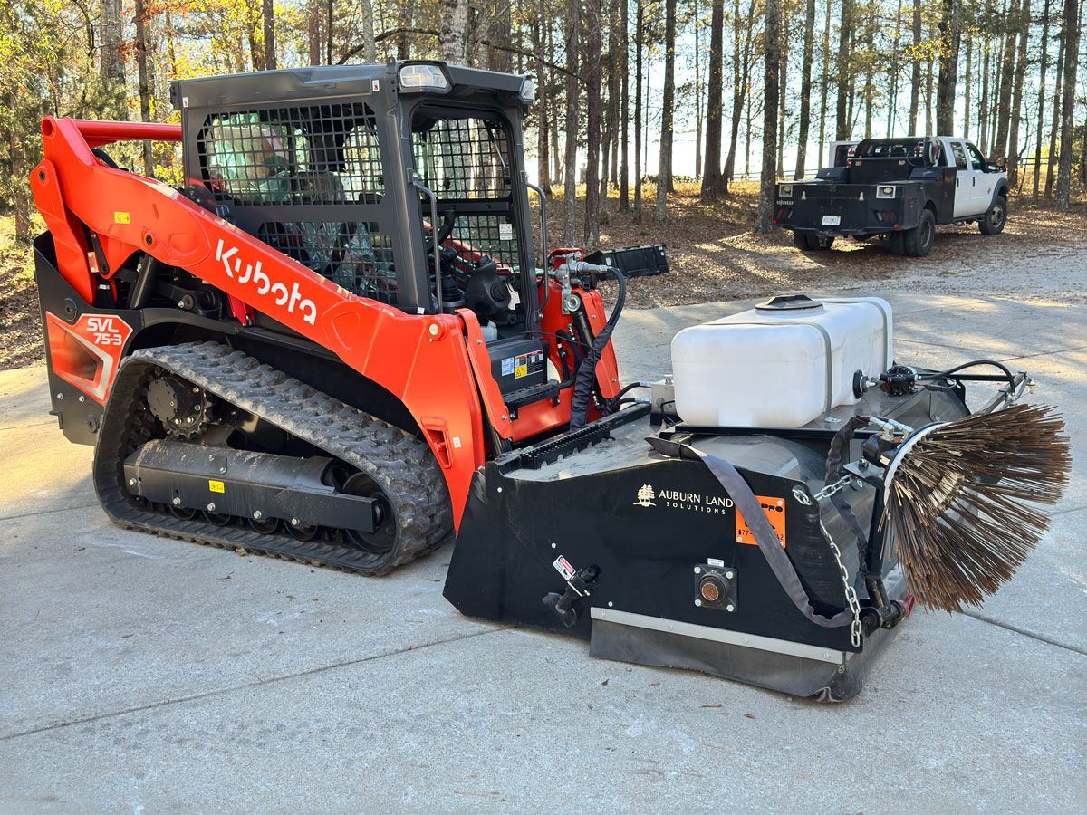 A red kubota skid steer with a broom attached to it is parked on the side of the road.