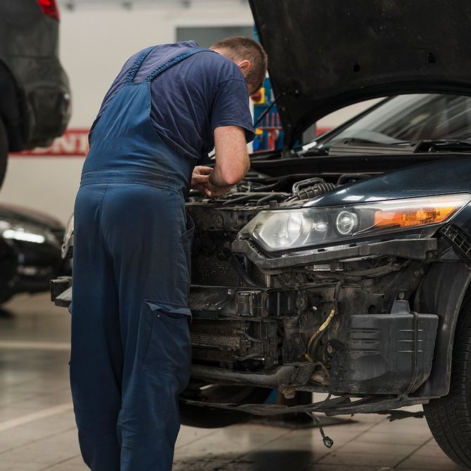 Mechanic in blue overalls working on a car with an open hood in a garage.