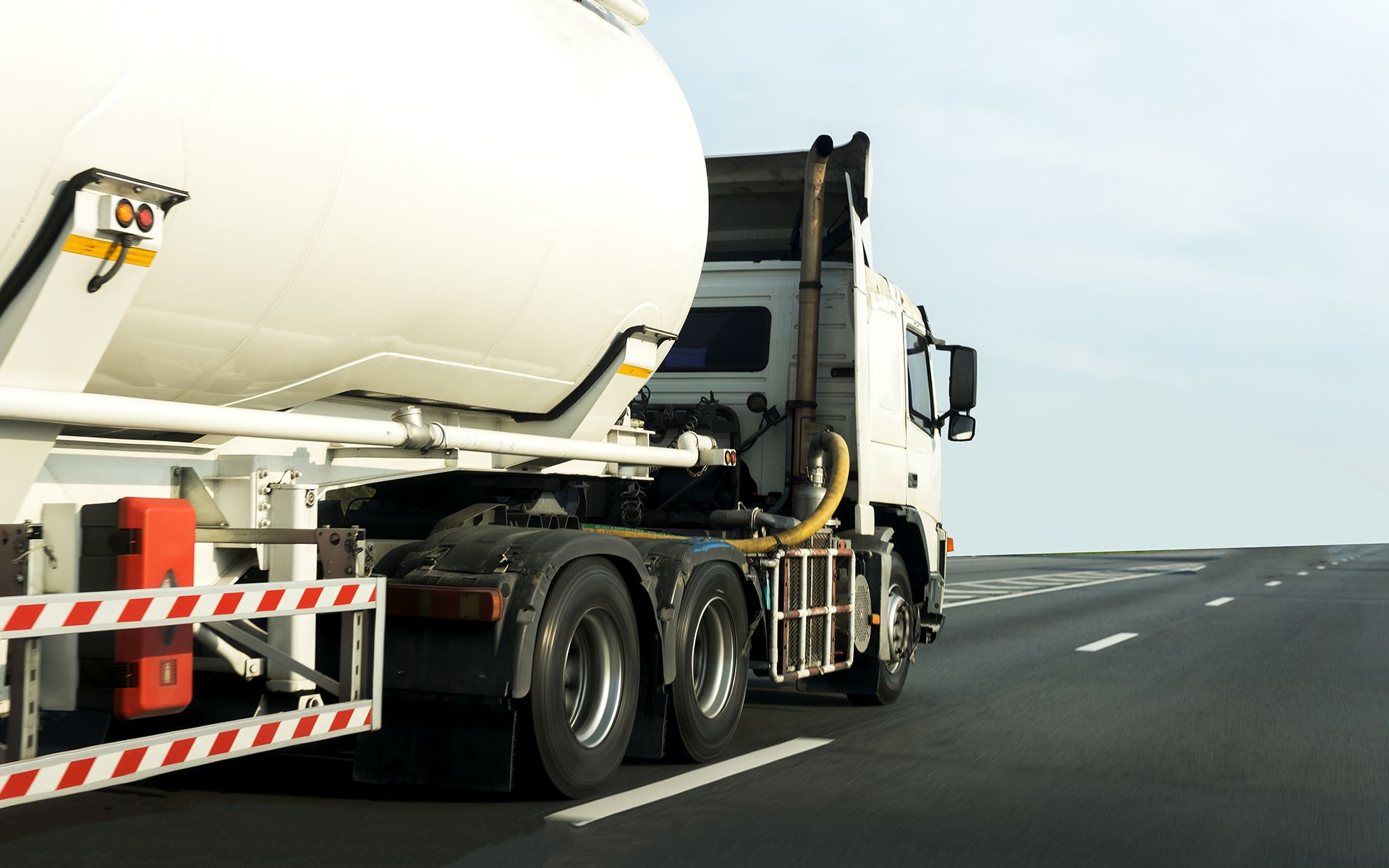 White tanker truck driving on an asphalt road under a light blue sky.