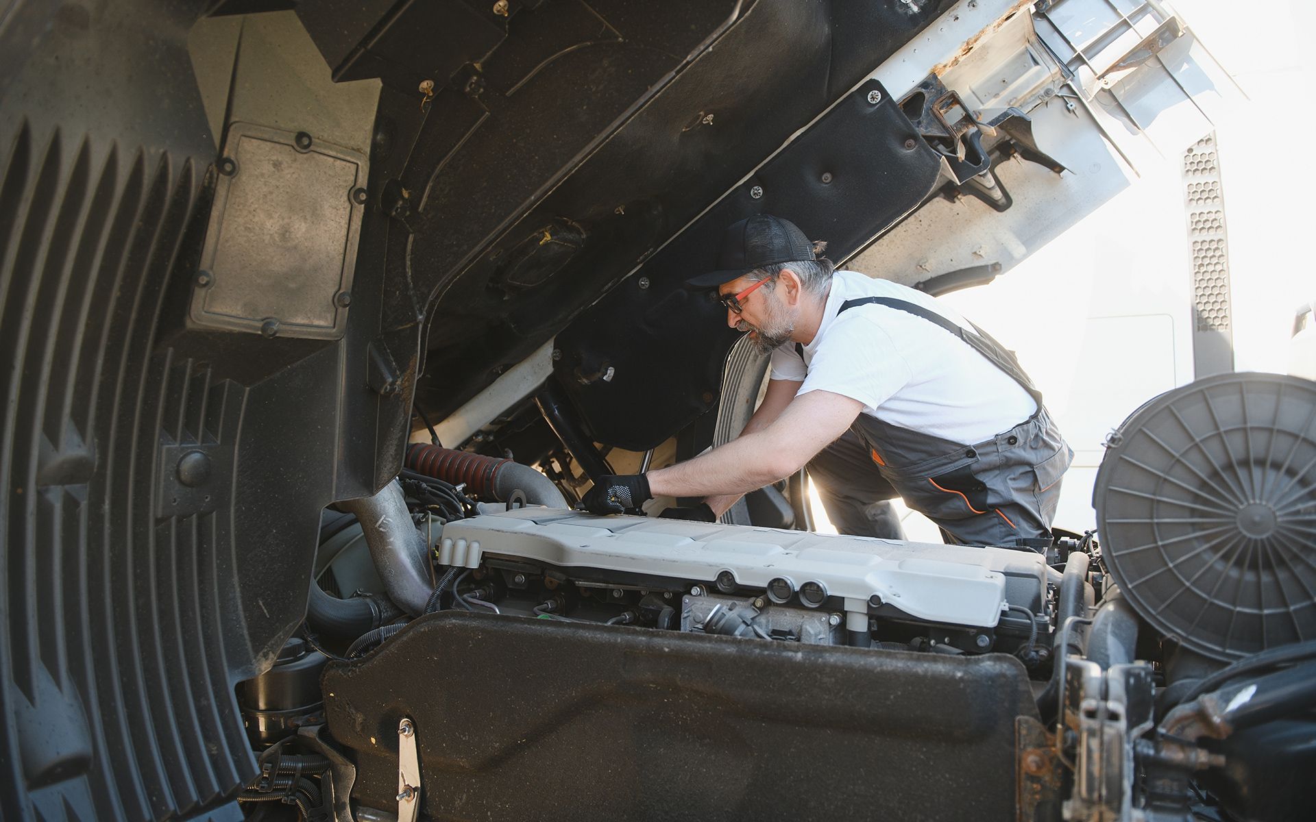 Mechanic in overalls working on a truck engine with the hood open on a sunny day.