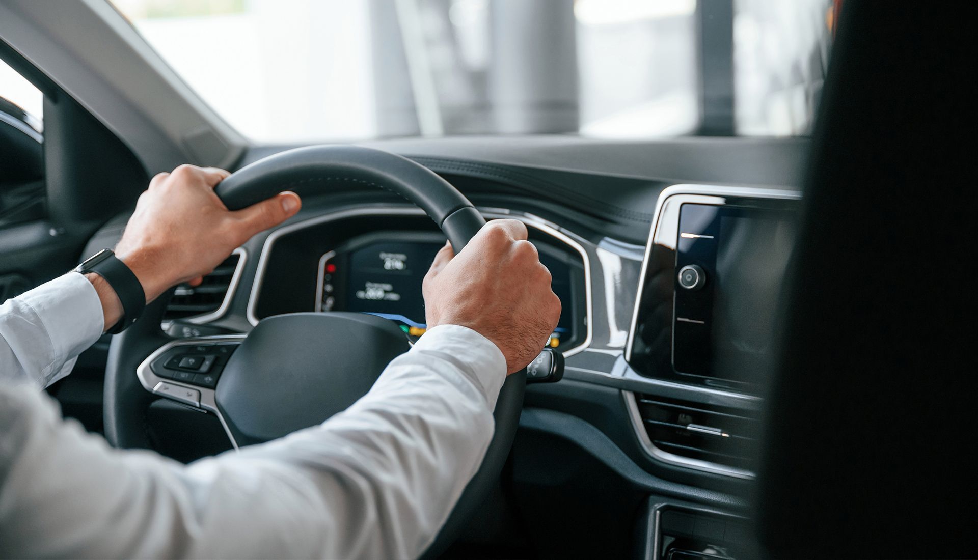 Person's hands on a car steering wheel in a showroom, wearing a white shirt.