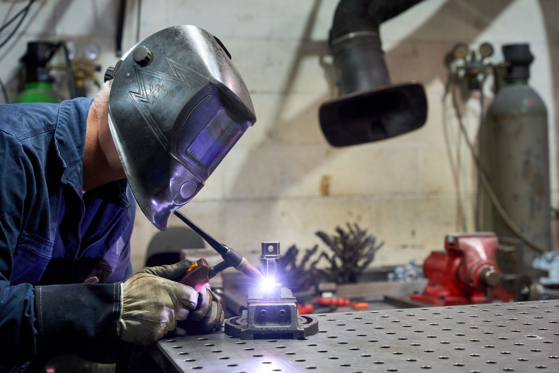 Welder in blue work clothes, welding a metal object with a bright arc in a workshop.