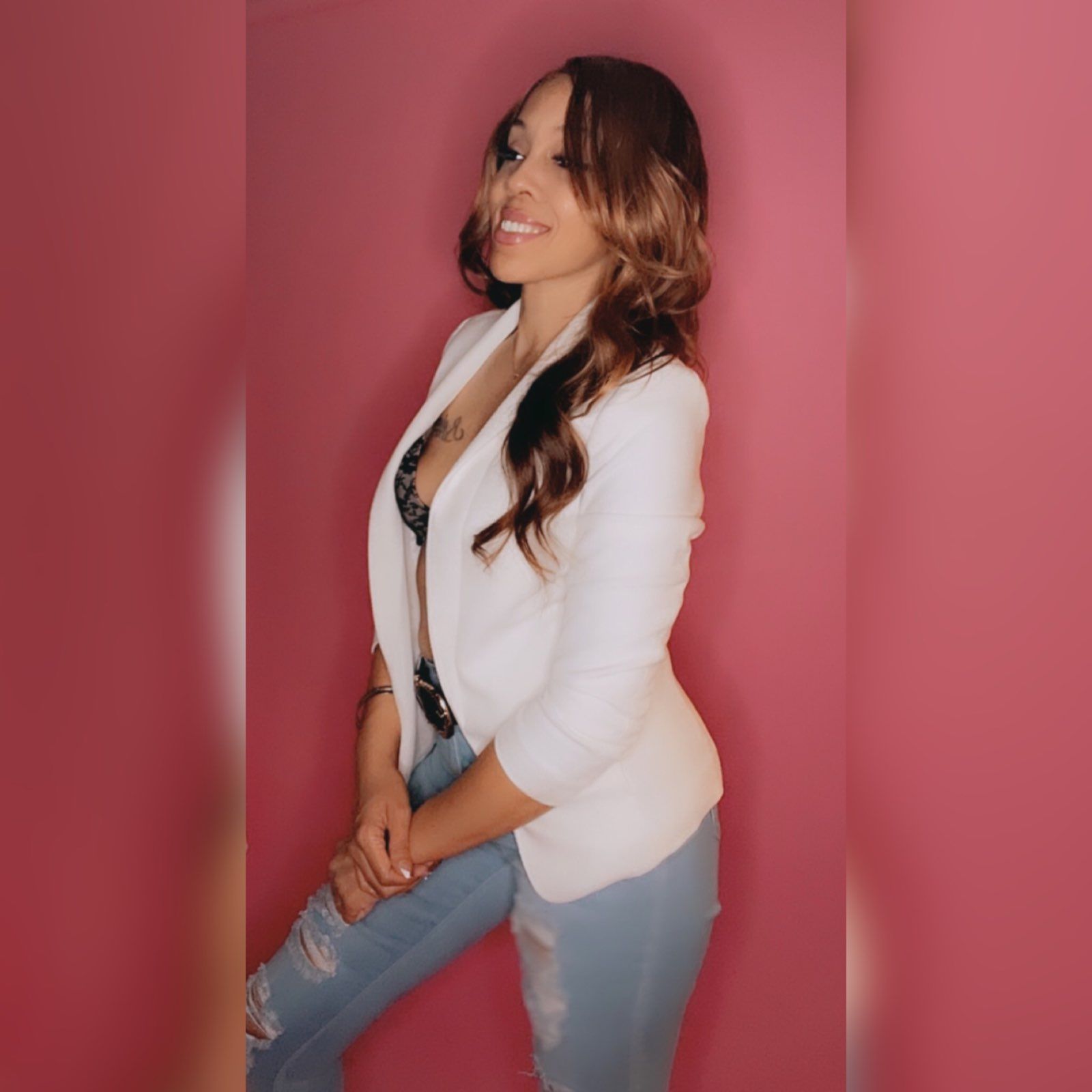 A woman in a white jacket and jeans is standing in front of a pink wall.