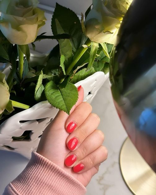 Una mano con smalto rosso acceso è tenuta vicino a un vaso di rose bianche sotto una luce calda.