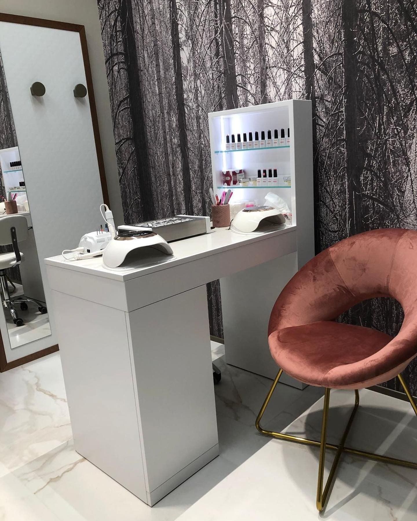 Una postazione per manicure bianca con un porta smalti e una sedia in velluto rosa su un pavimento di marmo, sullo sfondo di una carta da parati con motivo forestale.