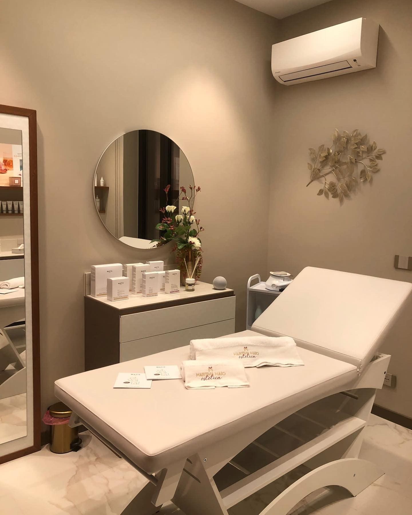 Una tranquilla sala trattamenti sui toni del beige, dotata di un lettino da massaggio professionale, uno specchio e prodotti estetici per la cura della pelle.