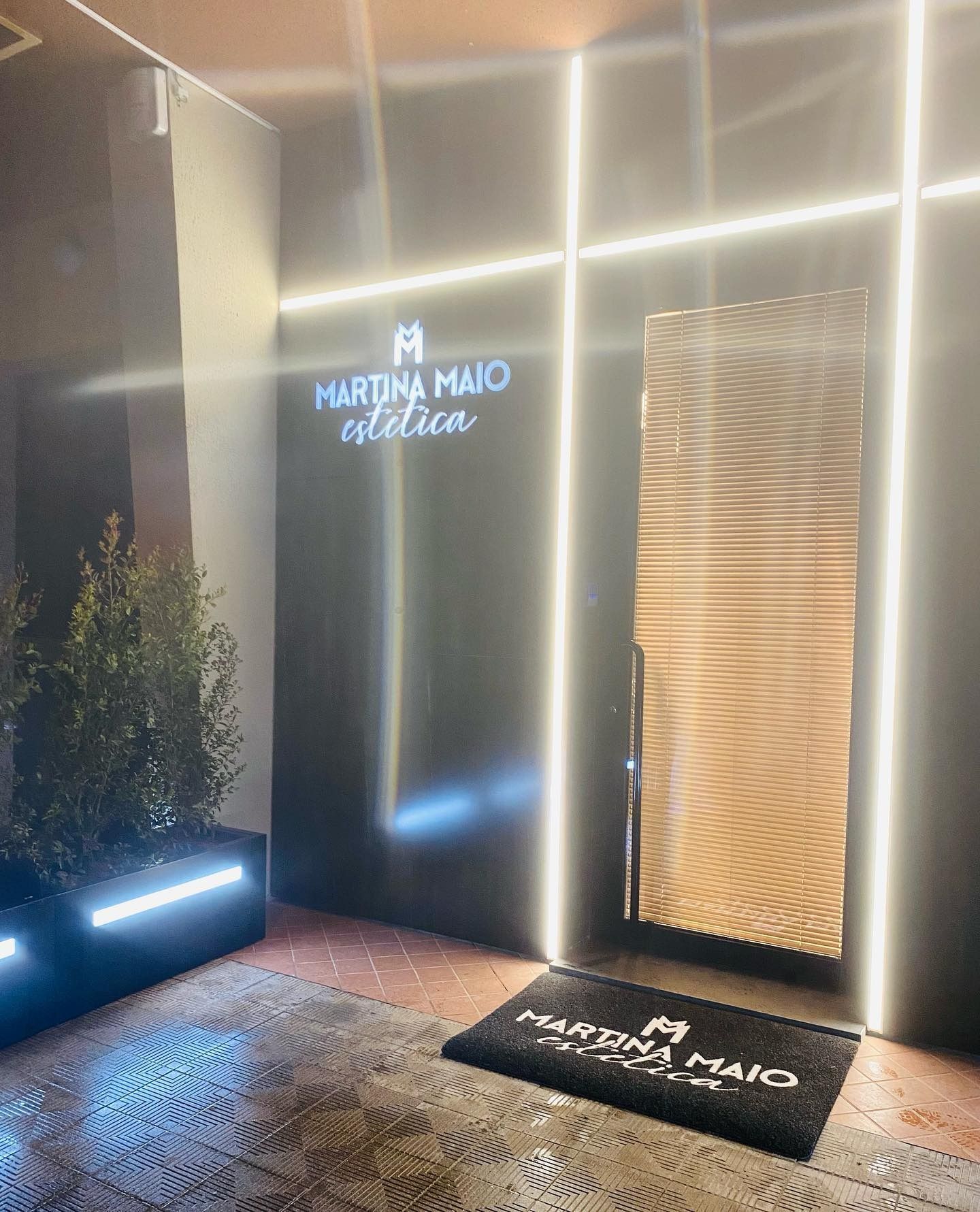 Ingresso illuminato del negozio "Martina Maio Estetica" con parete scura, strisce luminose verticali e zerbino con logo.