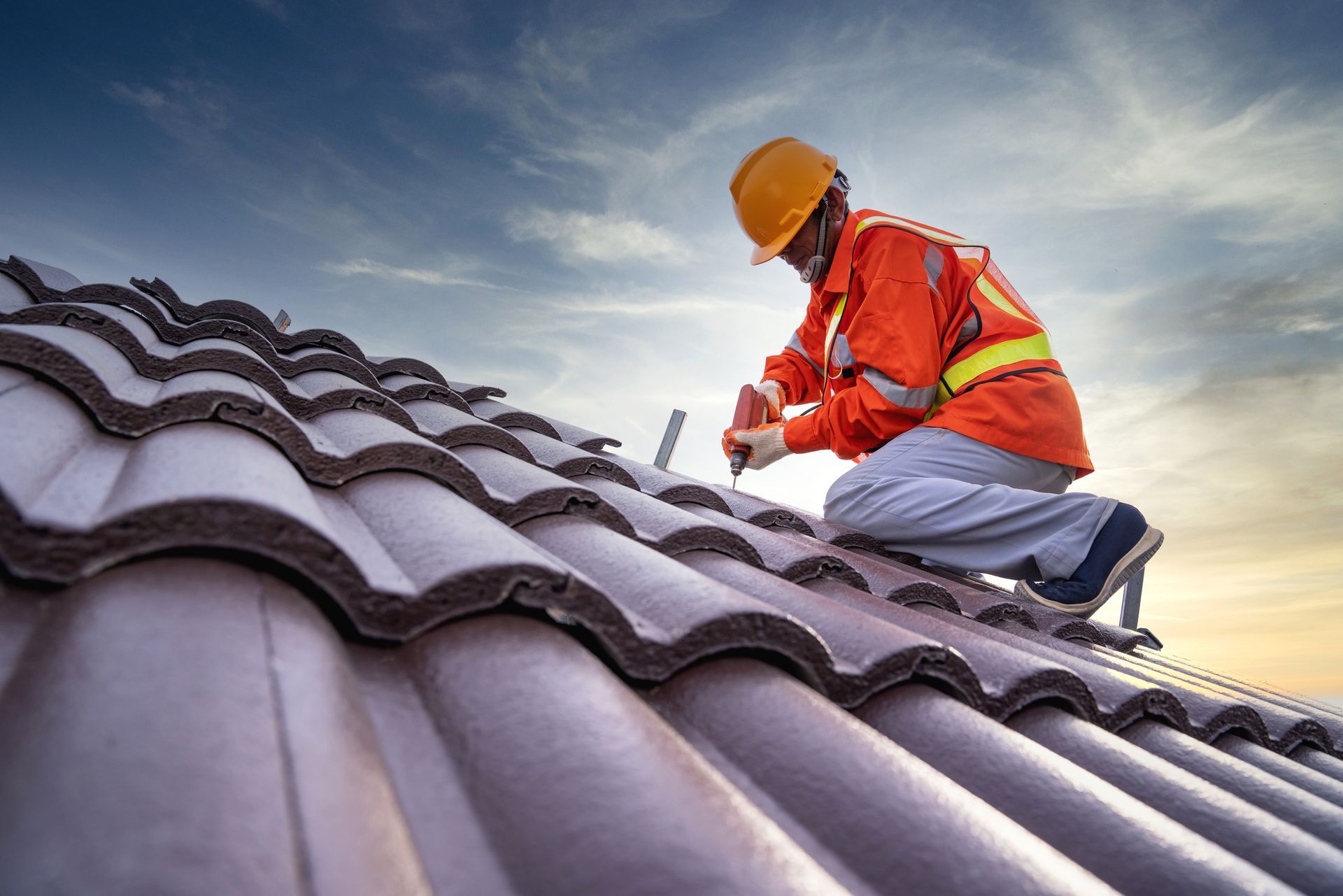 Construction worker installing roof tiles at sunset wearing safety gear and using power tools.