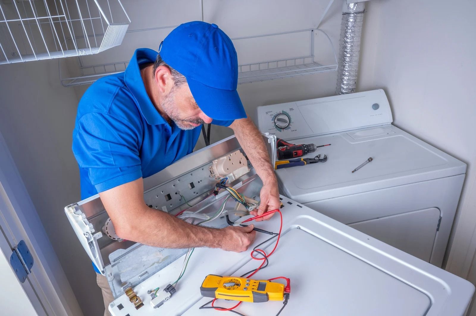 Man in blue shirt and cap repairing a white dryer with tools in a laundry room.