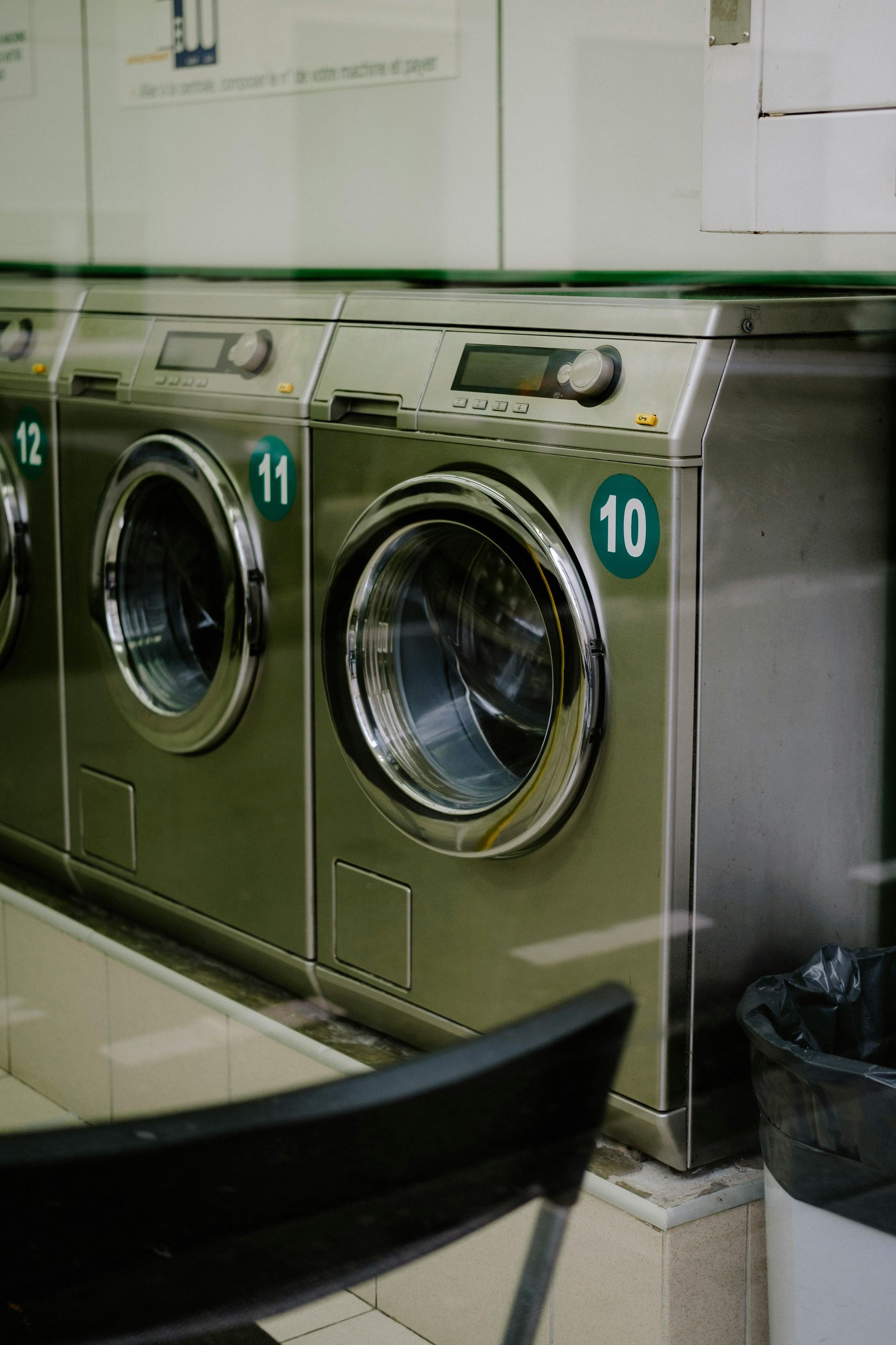 Row of stainless steel washing machines, numbered 10-12, in a laundromat. A black chair is in the foreground.