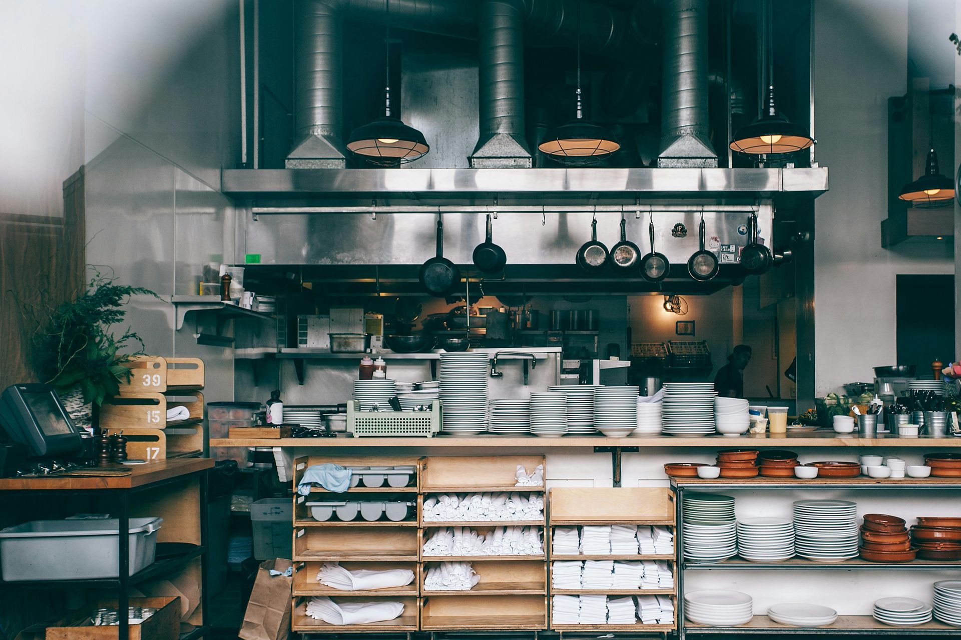 Restaurant kitchen with stainless steel hood, stacks of dishes, and hanging pots and pans.