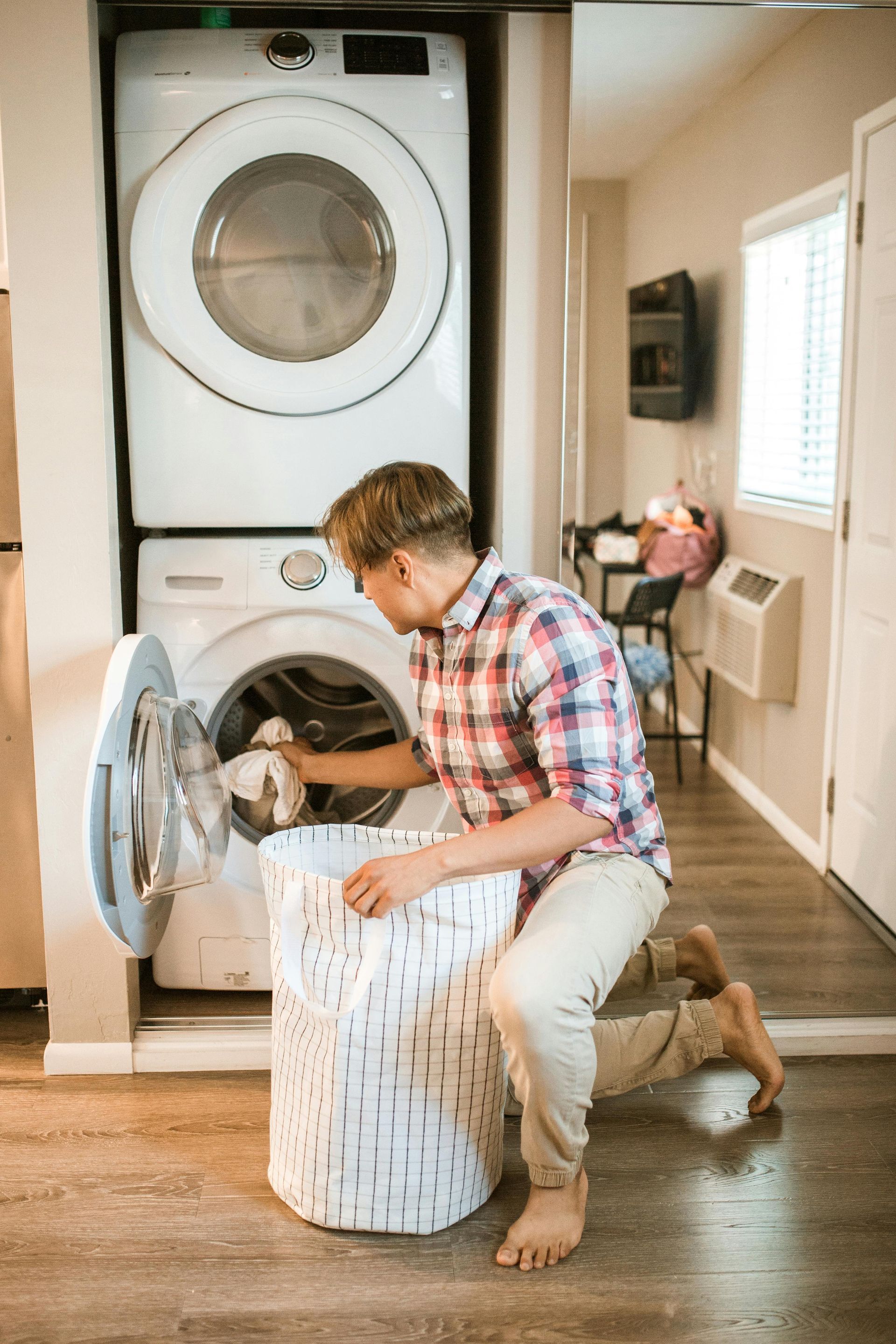 Man kneeling, putting clothes into a washing machine from a laundry basket in a laundry room.