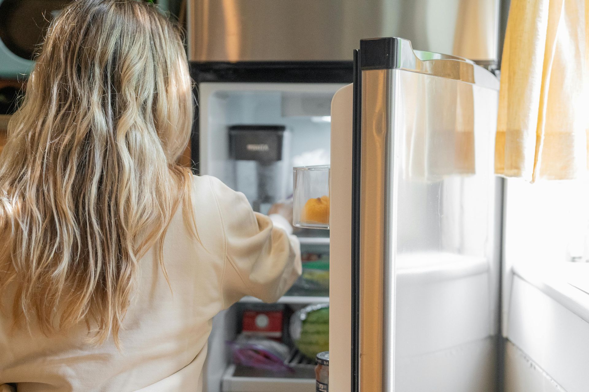 A person with shoulder-length blonde hair is reaching into an open stainless steel refrigerator in a bright kitchen.
