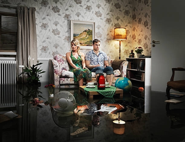Couple on sofa in flooded living room; water reflects furniture, lamp, and debris.