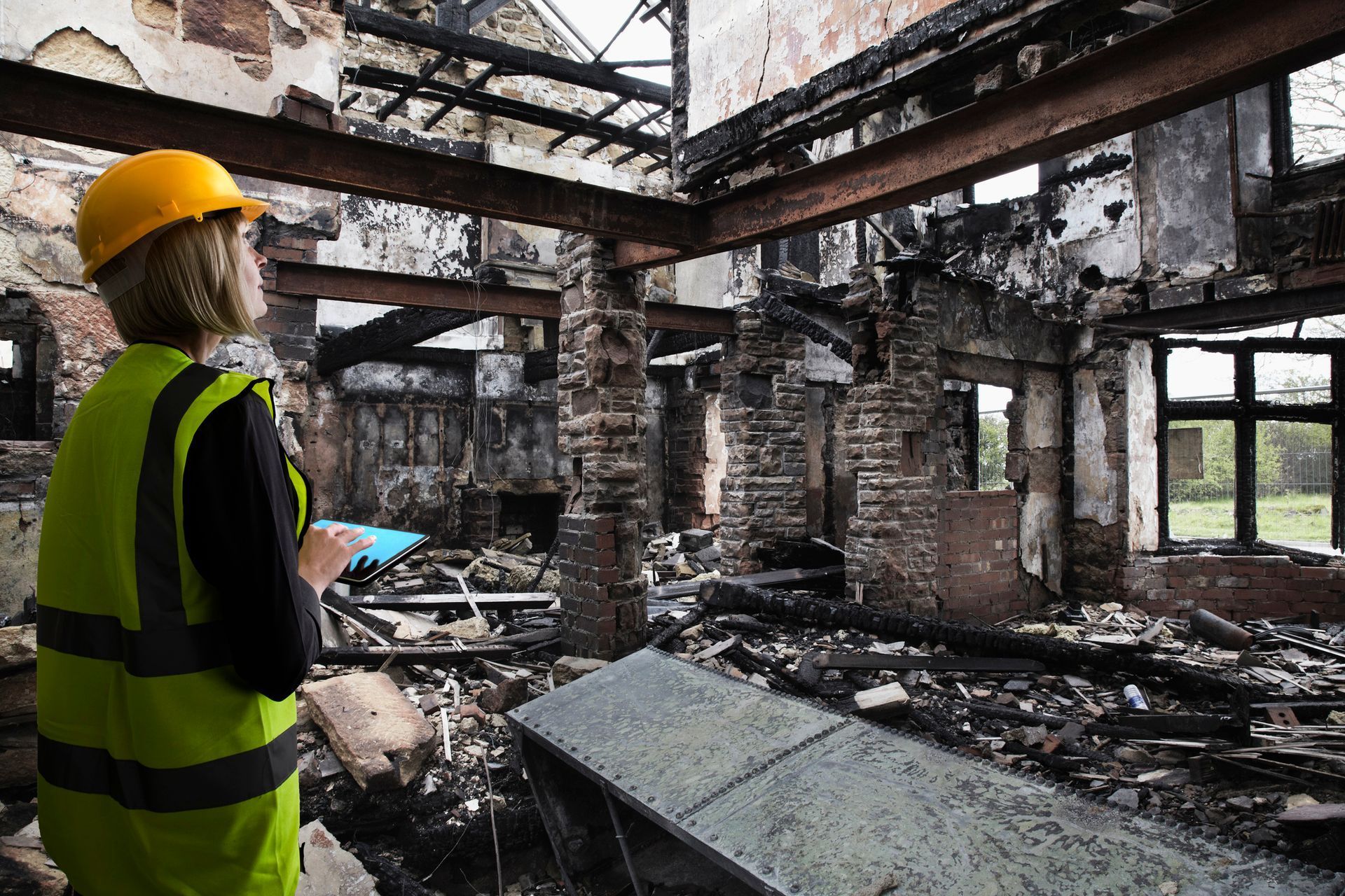 Person in hard hat and vest surveys fire-damaged building interior. Charred debris, exposed beams, and windows visible.