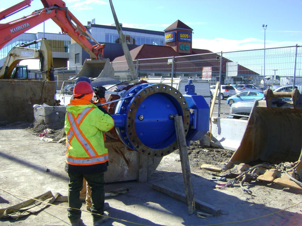 Construction worker in safety vest and helmet with a blue valve, excavator in background.