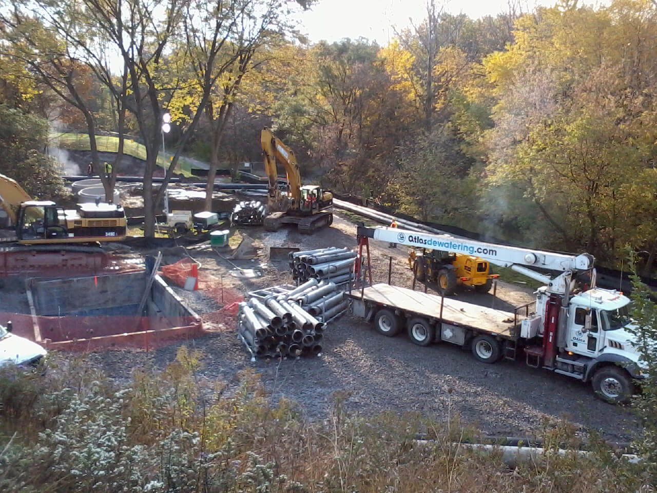 Construction site with heavy machinery and pipe bundles, autumn foliage in the background.