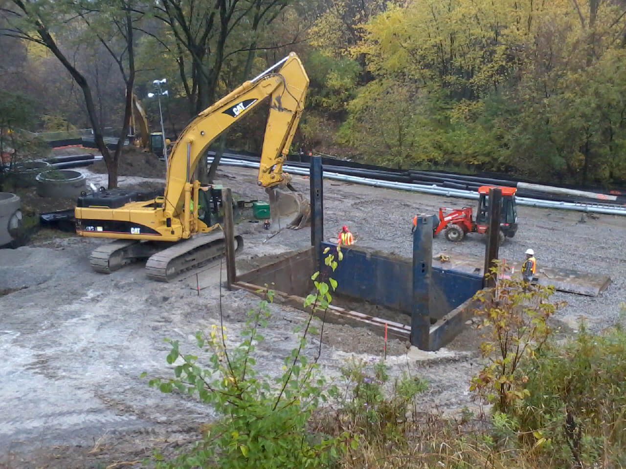 Construction site: Excavator digging trench, workers, pipes, and a small loader.