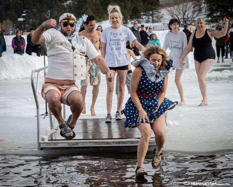 People jumping into icy water from a dock. Woman in polka dot dress reacts, others smile. Snowy outdoor setting.
