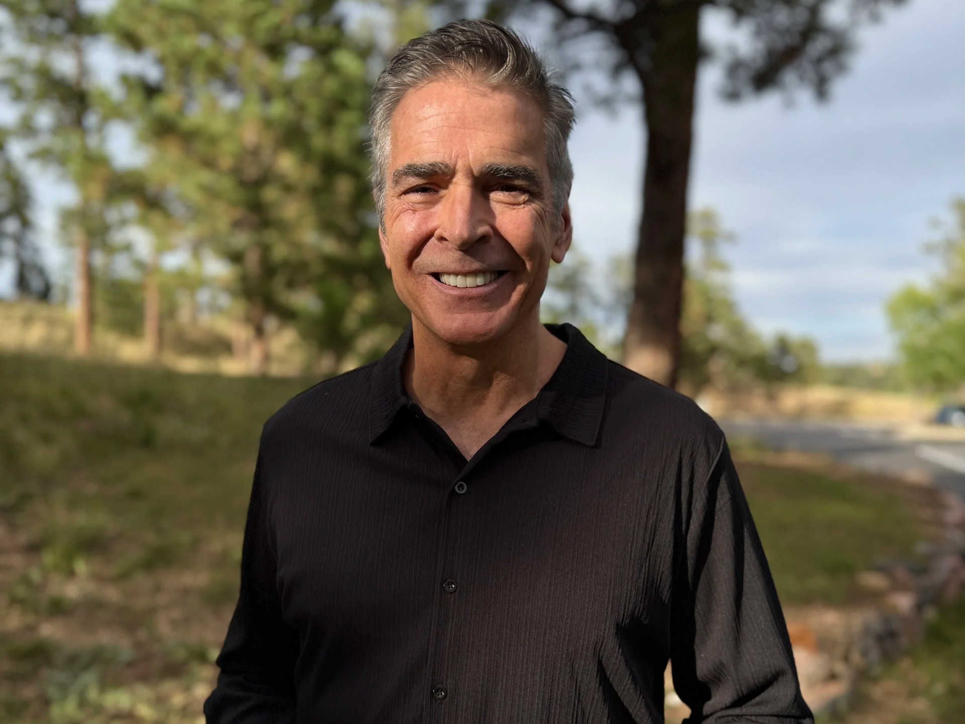 Man with graying hair smiles outdoors, wearing a black collared shirt in a sunny field.