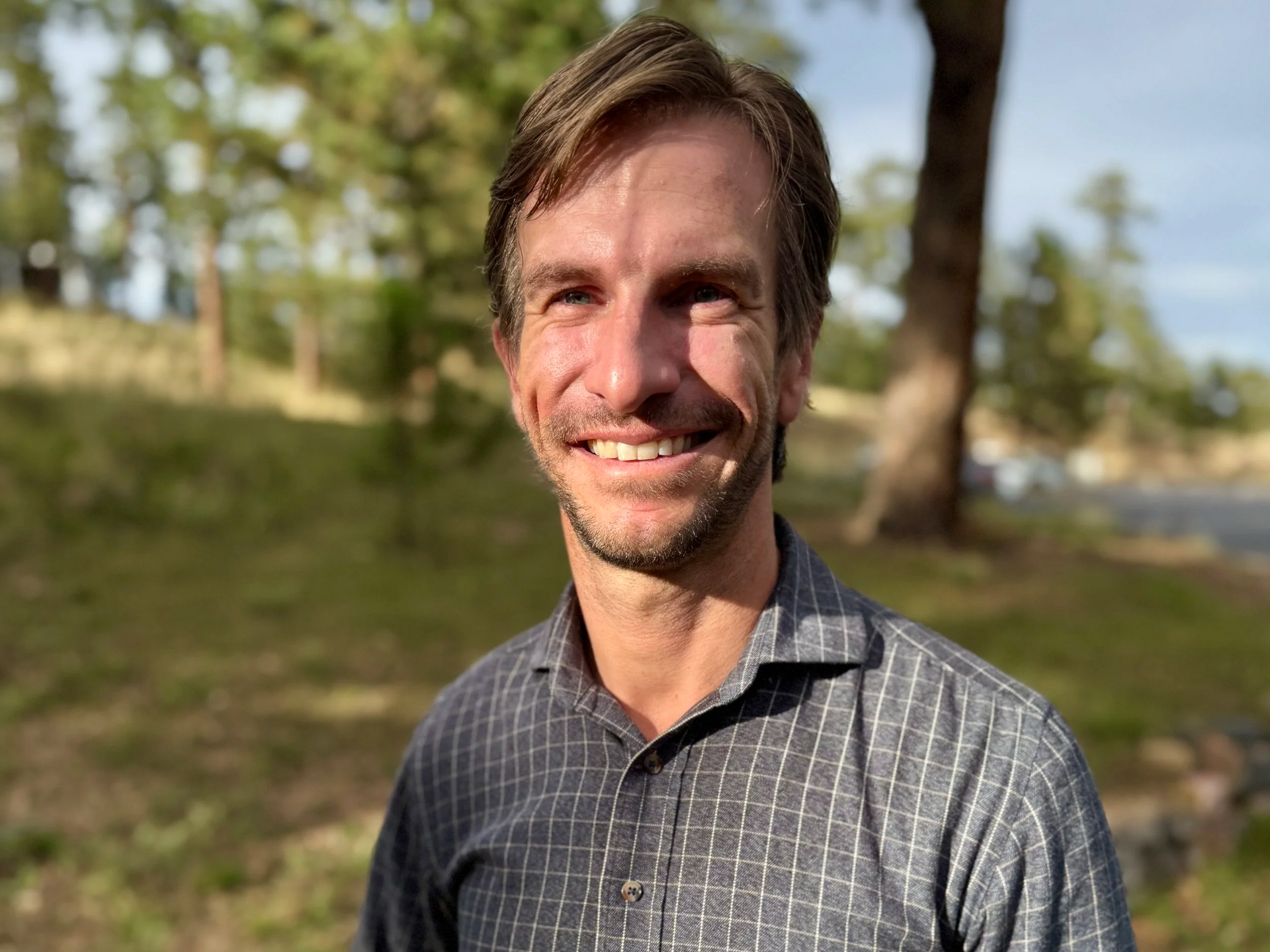 Man with a light smile in a gray plaid shirt outside, trees in the blurred background.