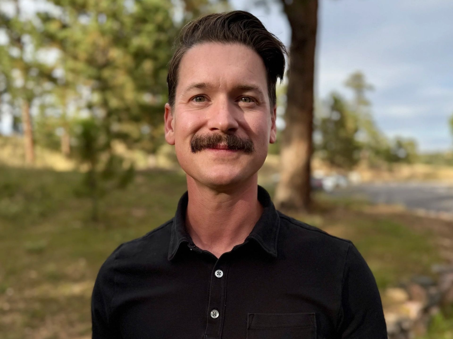 Man with dark mustache and black shirt outdoors, smiling.