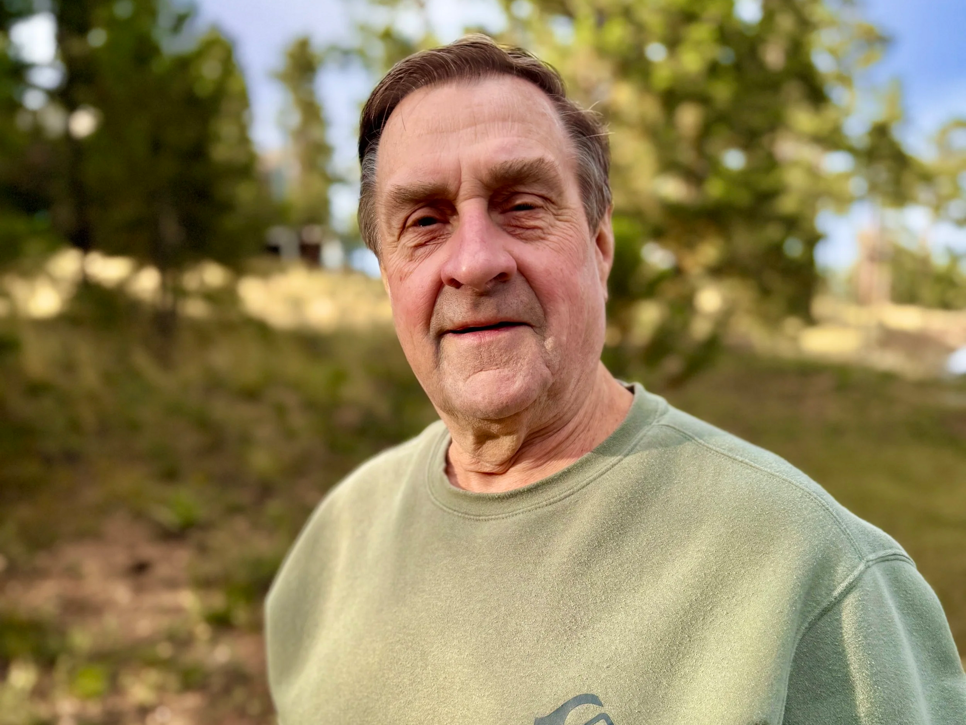 Man in green shirt smiles outdoors, with trees in the background.