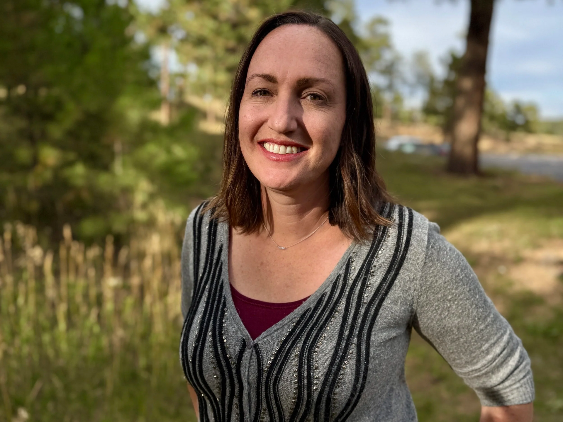 Woman smiling outdoors, wearing a gray top with black detailing and a burgundy undershirt, with trees and grass in the background.