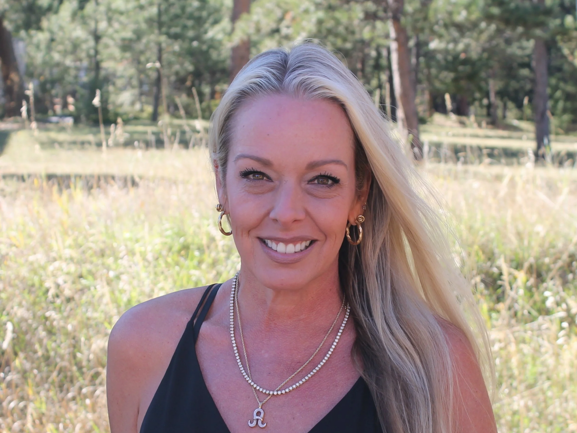 Woman with blonde hair wearing a necklace and hoop earrings, smiling outdoors with trees in the background.