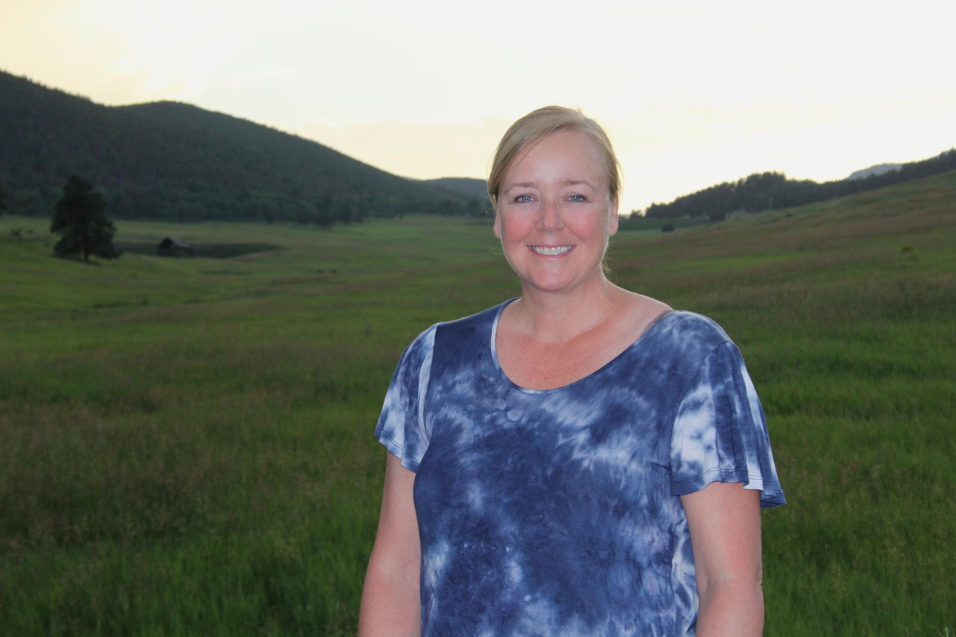 Woman smiles in front of a green field, mountains in the background, wearing a blue tie-dye shirt.