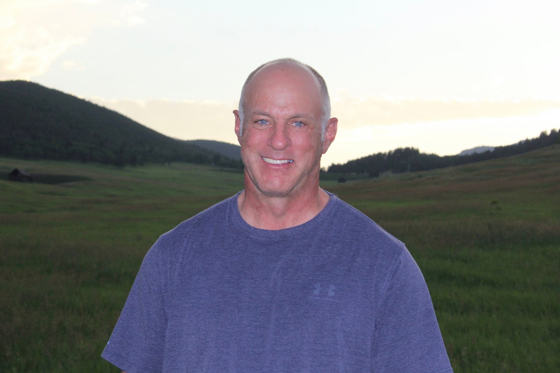 Man smiling outdoors in front of a green field and hills, wearing a blue shirt.