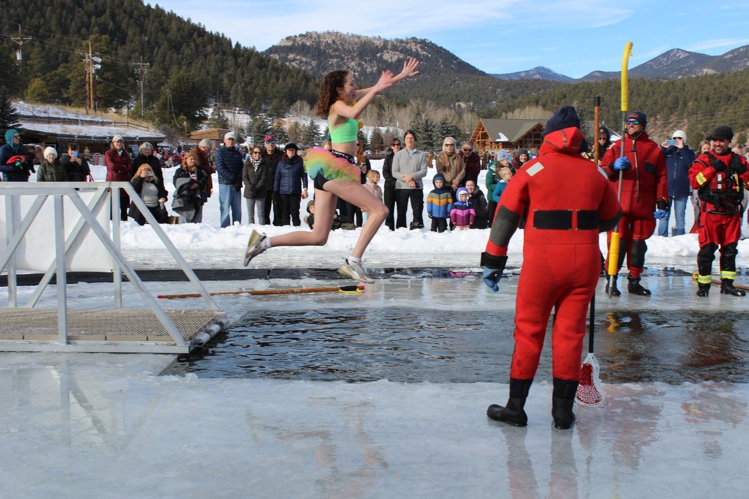 Person leaps into icy water at an outdoor event, cheering, with rescuers and onlookers in a snowy landscape.