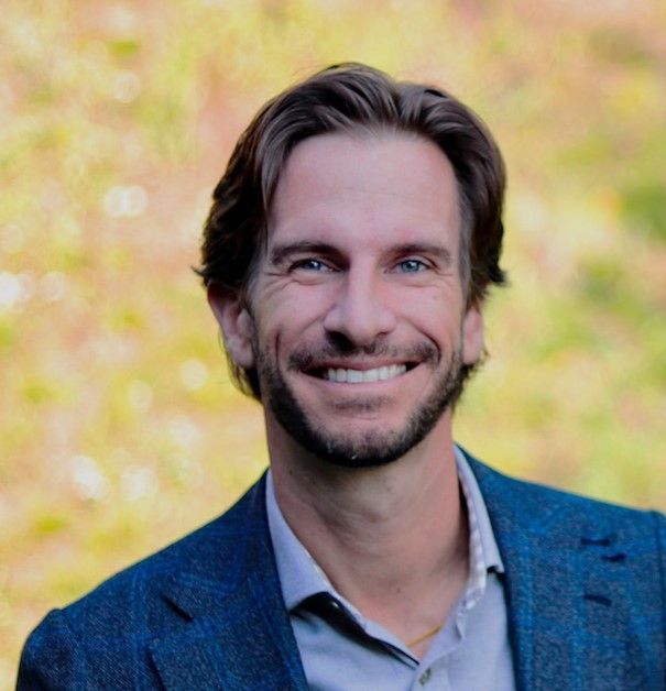 Man with a light smile in a gray plaid shirt outside, trees in the blurred background.