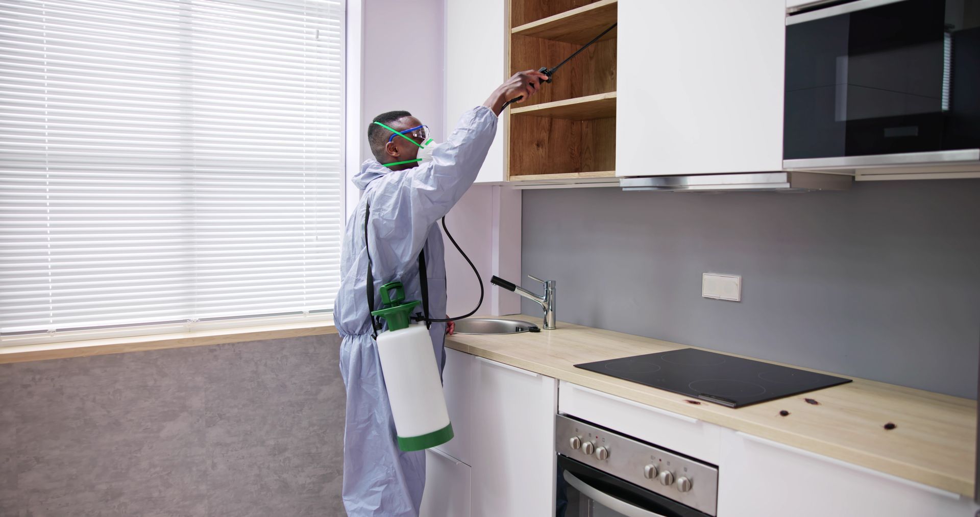 Person in protective suit spraying kitchen cabinets with insecticide.